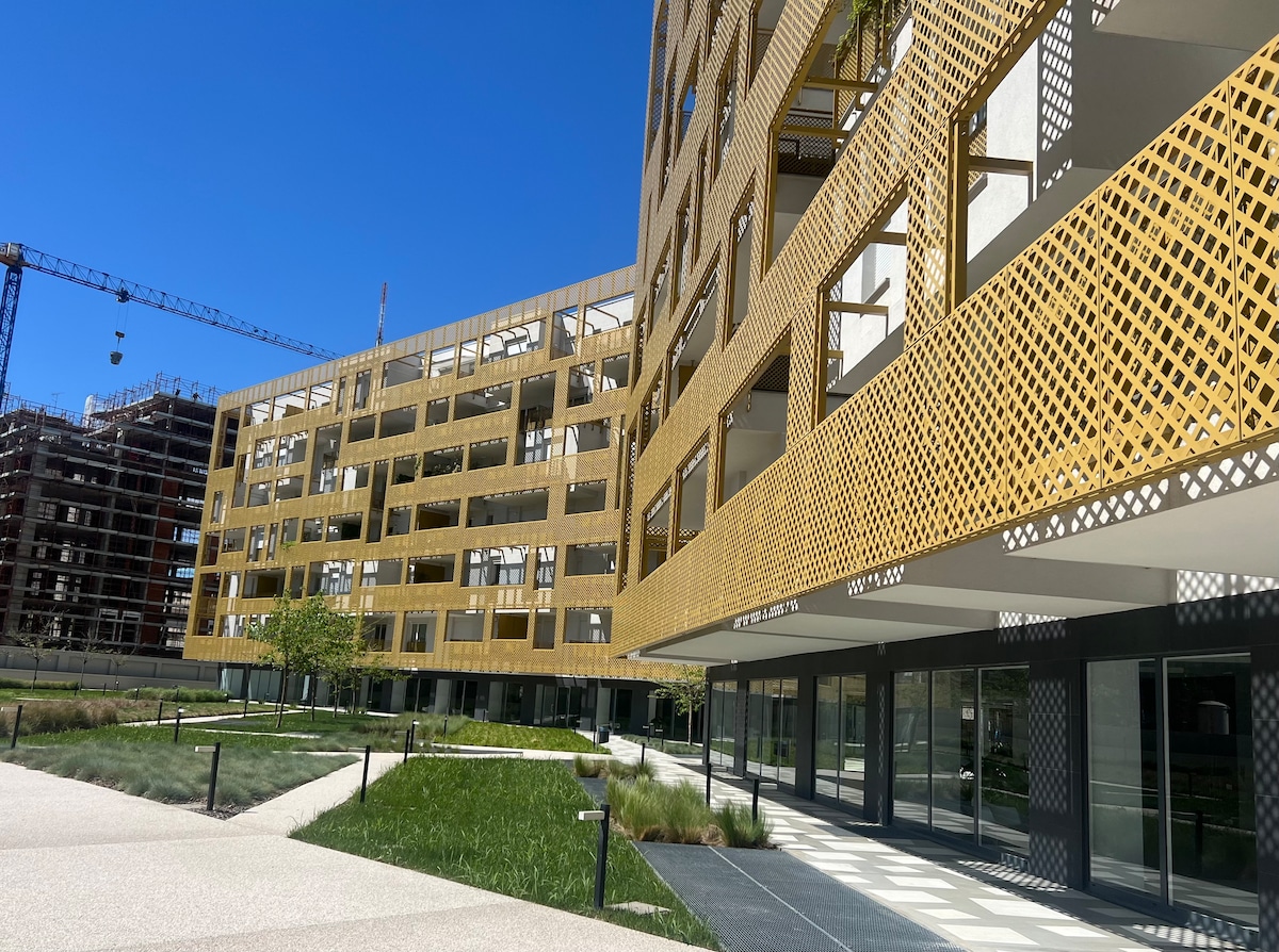 The image presents a modern residential complex featuring distinctive golden lattice work on the exterior. Lush green landscaping and walkways are visible in the foreground, complemented by a clear blue sky overhead. Construction activity is noted in the background.