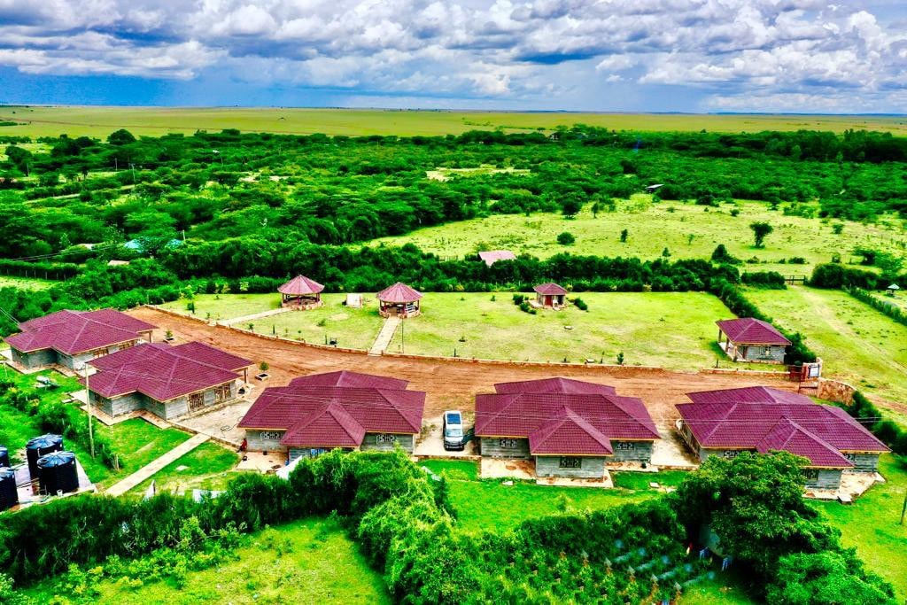 An aerial view showcases a collection of cottages arranged around a central pathway. Each cottage features a red roof and is surrounded by lush green landscaping. The expansive surroundings include open fields and patches of trees, under a cloudy sky.