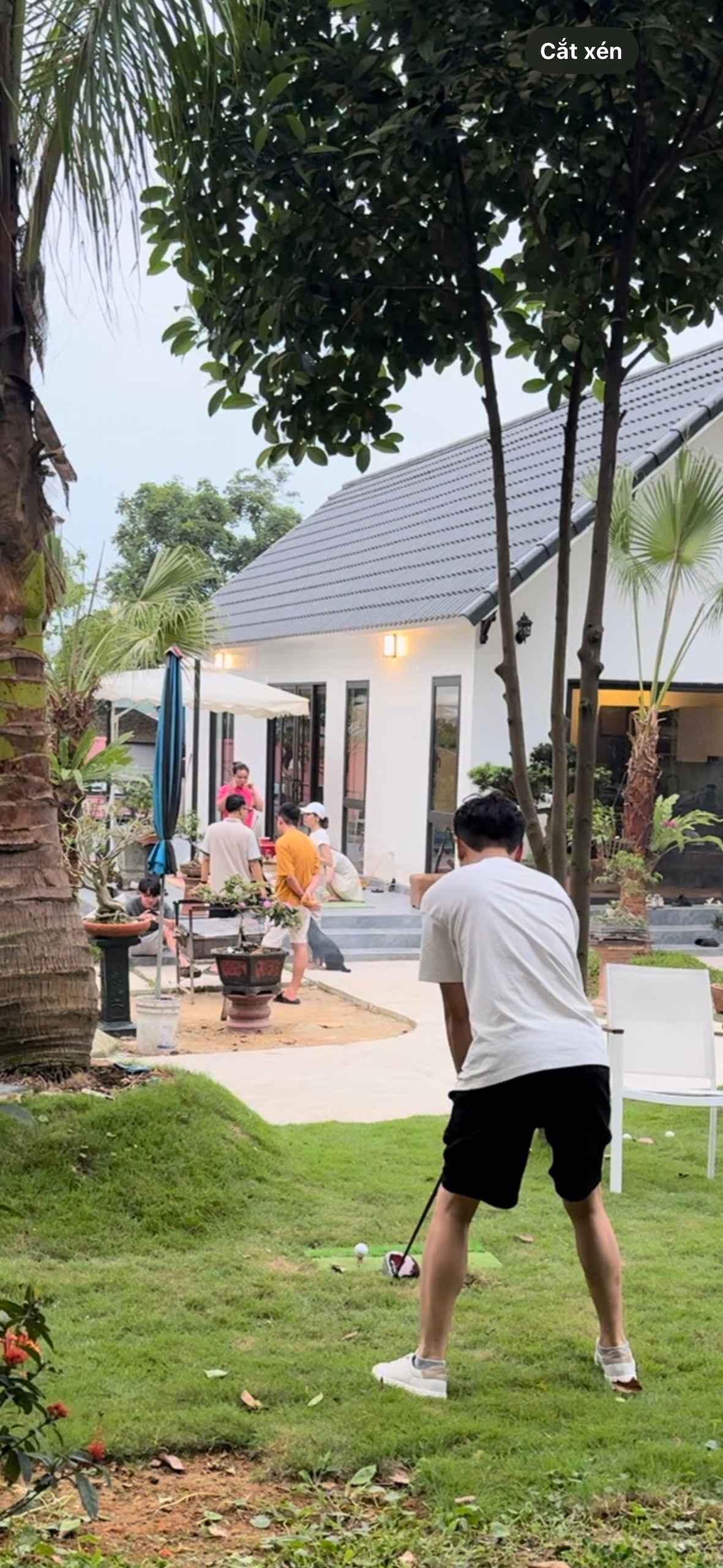 A man is seen preparing to swing a golf club on a neatly maintained lawn bordered by tropical plants. In the background, a modern villa with large glass doors and outdoor seating is visible, along with a swimming pool and guests enjoying the setting.