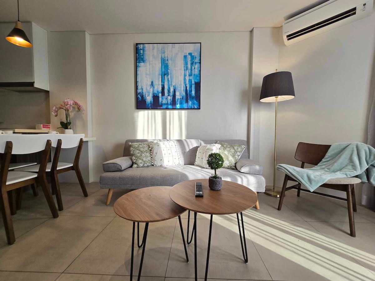 A modern living space is presented with a cozy gray sofa adorned with patterned cushions. Two small wooden tables are positioned in front, while a stylish floor lamp and artwork add interest. A dining table is visible in the background, surrounded by four chairs.