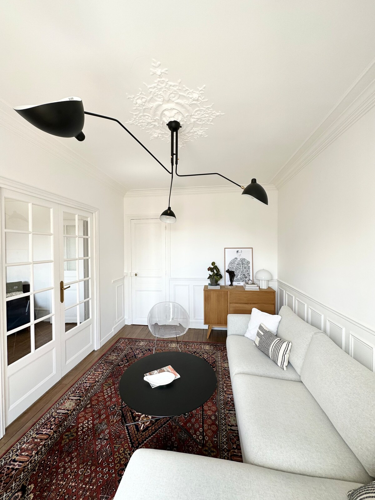 A bright living room area features a light grey sofa positioned against a wall adorned with detailed molding. A round black table sits in front, while a chic floor lamp with two arms provides illumination. Glass-paneled doors lead to an adjoining space.