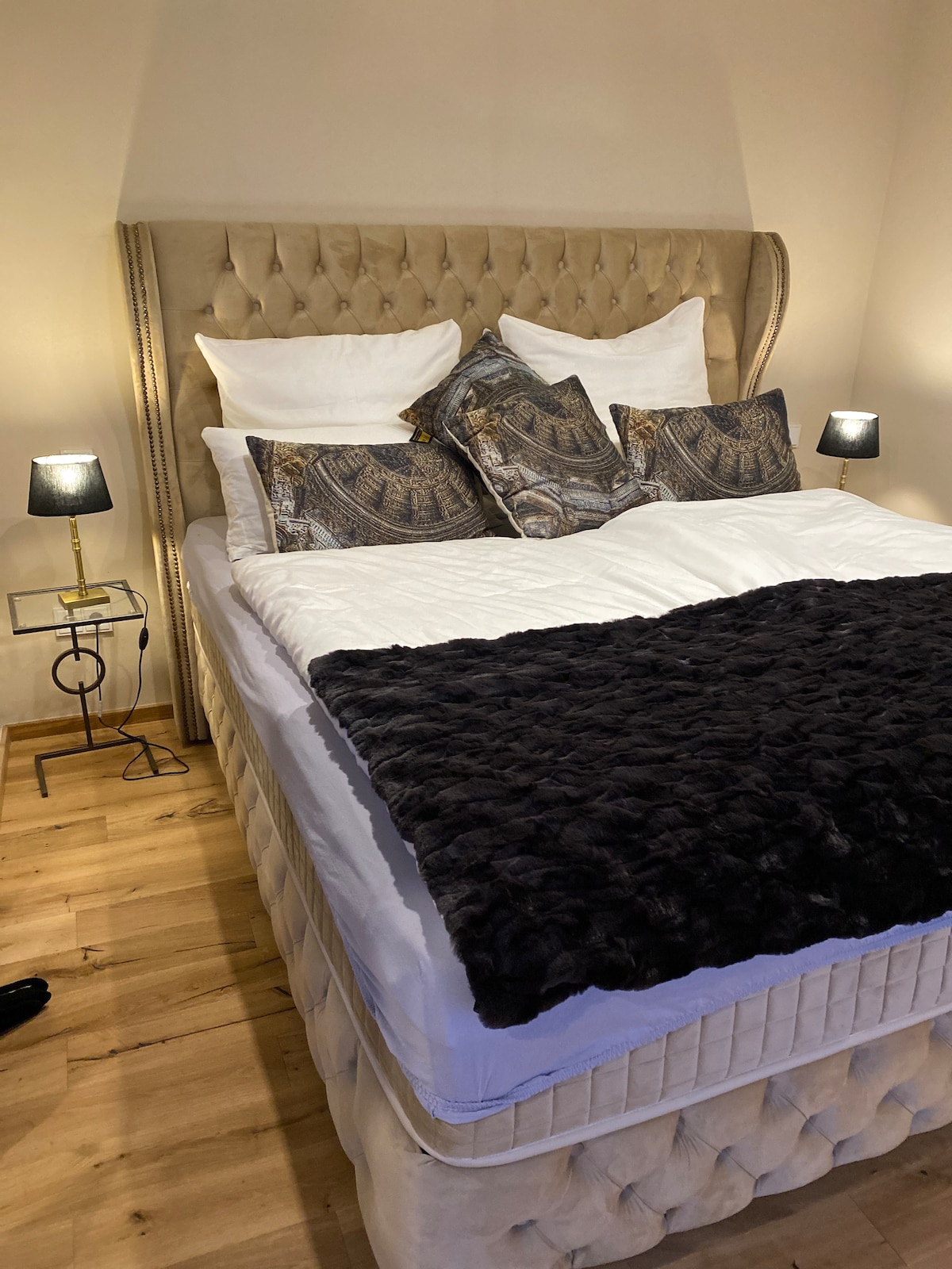 A spacious bed with a tufted headboard is dressed in crisp white linens, accompanied by decorative pillows featuring intricate patterns. A soft, dark throw blanket is layered across the bed, enhancing the inviting appearance. Bedside lamps provide soft lighting, illuminating the warm wooden floor.