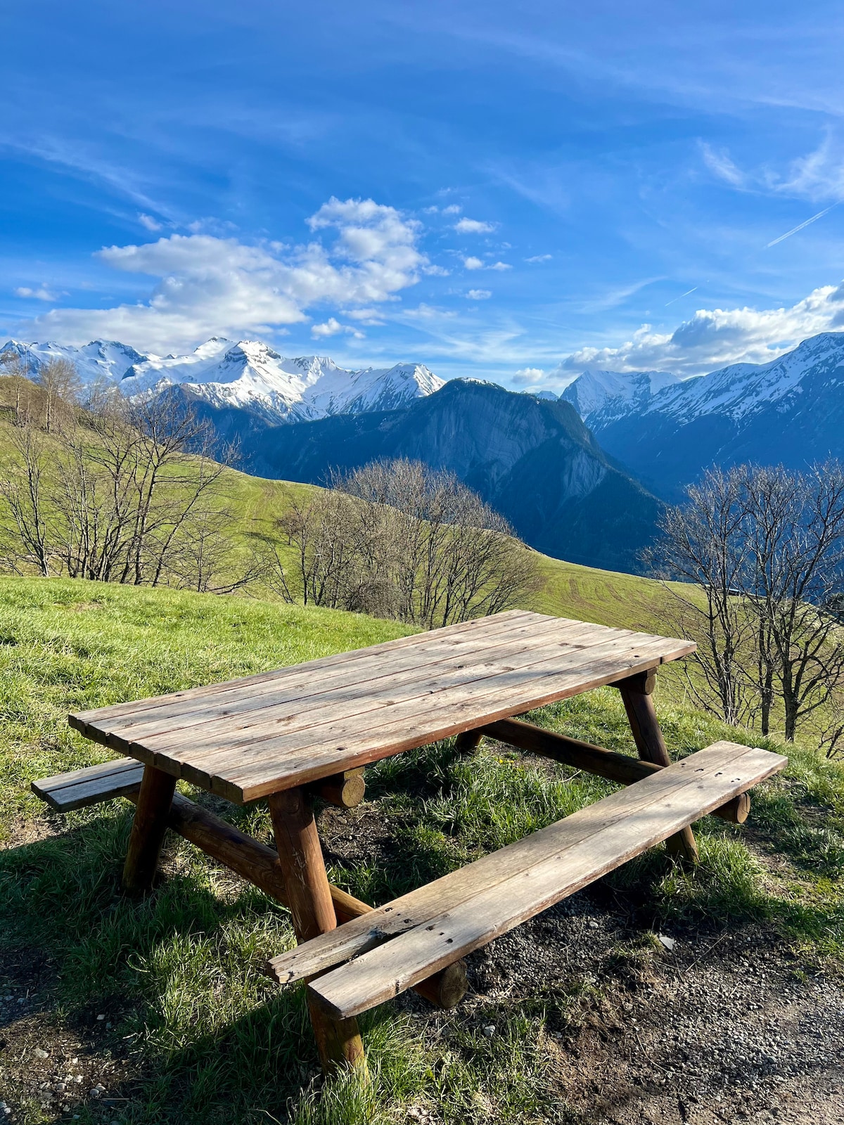 An outdoor wooden picnic table with attached benches is positioned on a grassy hillside, offering sweeping views of surrounding mountains. The landscape features a mix of snow-capped peaks and lush greenery under a bright blue sky, creating an inviting setting for relaxation.