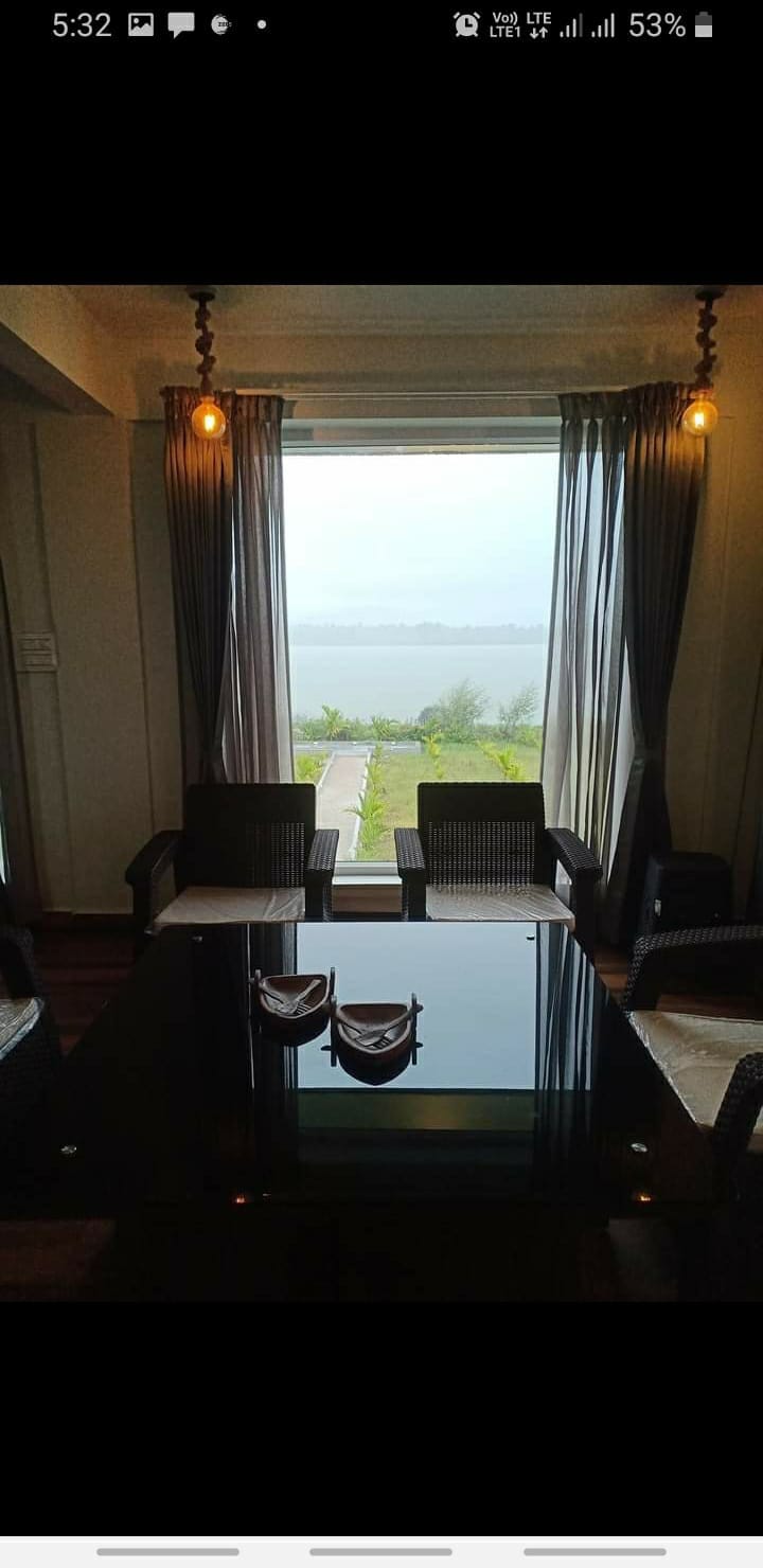 A cozy seating area is presented with two woven chairs and a dark glass table. Floor-to-ceiling windows allow natural light to flood the space, featuring views of the tranquil lake and surrounding greenery. Soft curtains frame the window, adding a touch of warmth.
