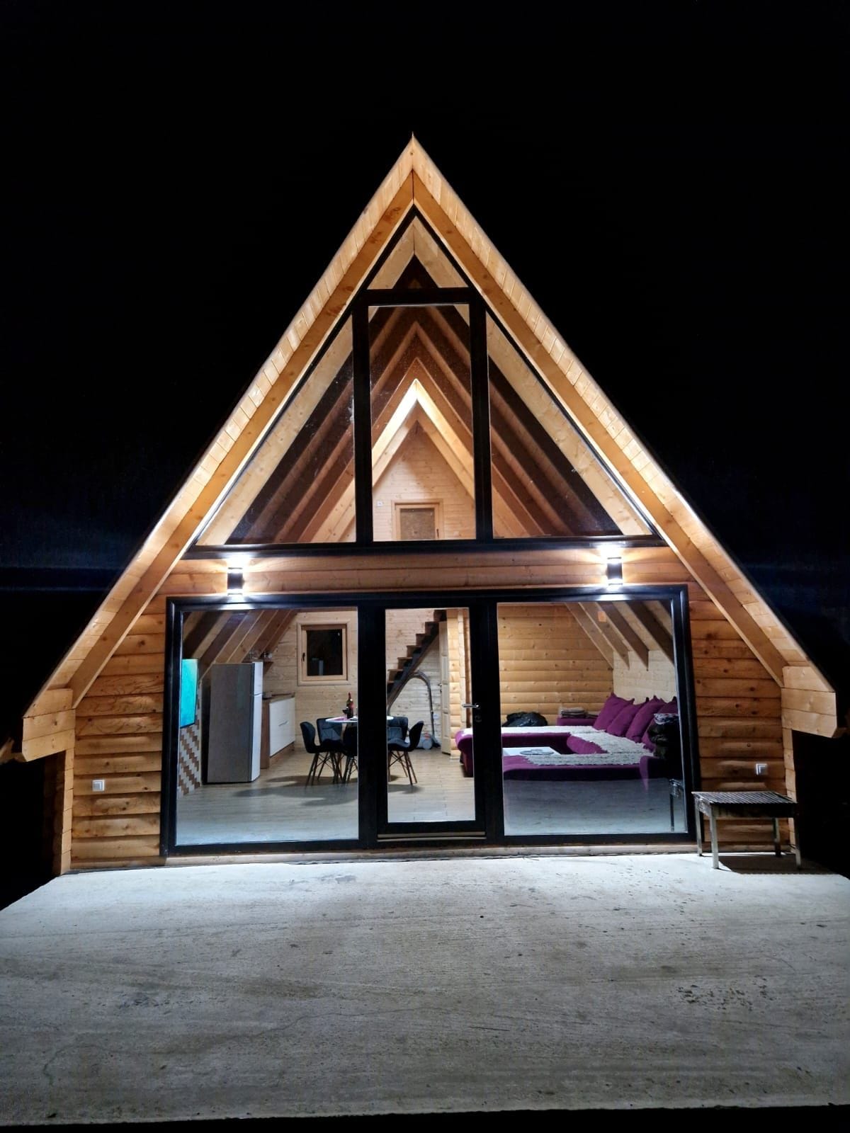 The exterior of a wooden chalet is showcased at night, featuring an A-frame design. Bright lighting emphasizes the intricate woodwork and spacious entrance, with a view into the warmly lit interior that includes a dining area and seating. The surrounding area is dark, enhancing the cabin's inviting glow.