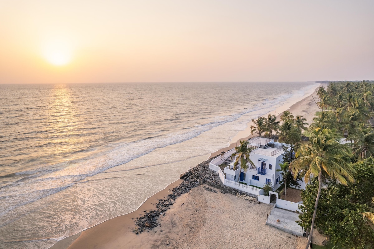 A cozy villa sits along a serene beach during sunset, reflecting warm tones as the sun descends over the horizon. Gentle waves lap against the sandy shore, framed by swaying palm trees, creating a tranquil coastal scene.