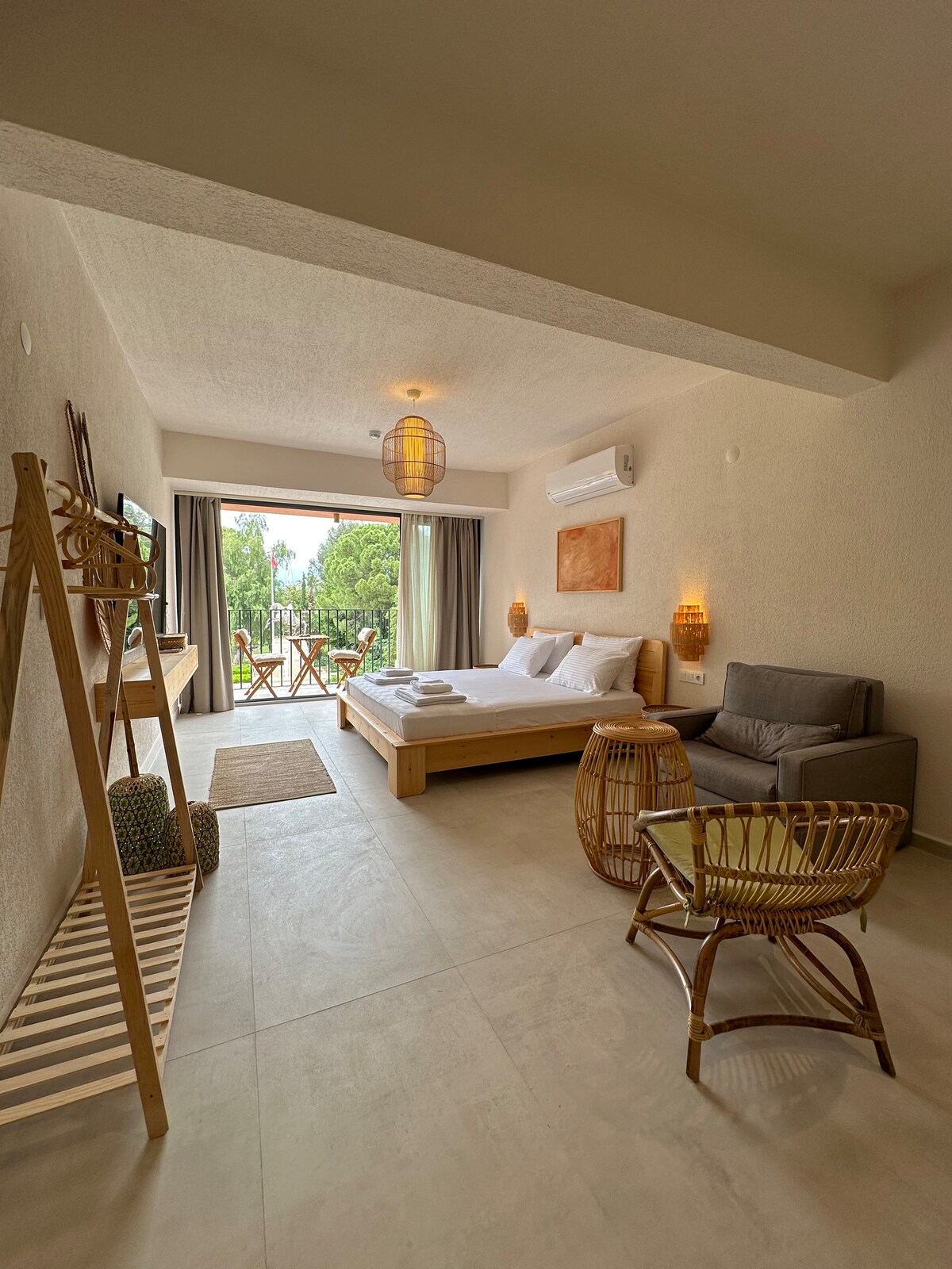 A light-filled room features a spacious bed with neatly arranged linens, complemented by a cozy seating area with a grey armchair and a round wooden table. Large windows offer views of greenery, while woven decor elements enhance the tranquil ambiance.