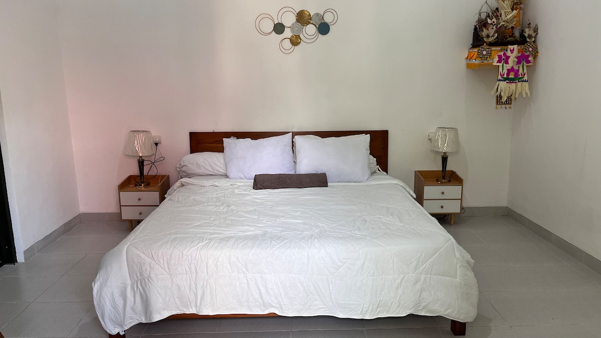 A spacious bed is positioned centrally against a white wall, adorned with decorative wall art that features circular elements in various colors. Two bedside tables with lamps flank the bed, creating a balanced and serene environment.