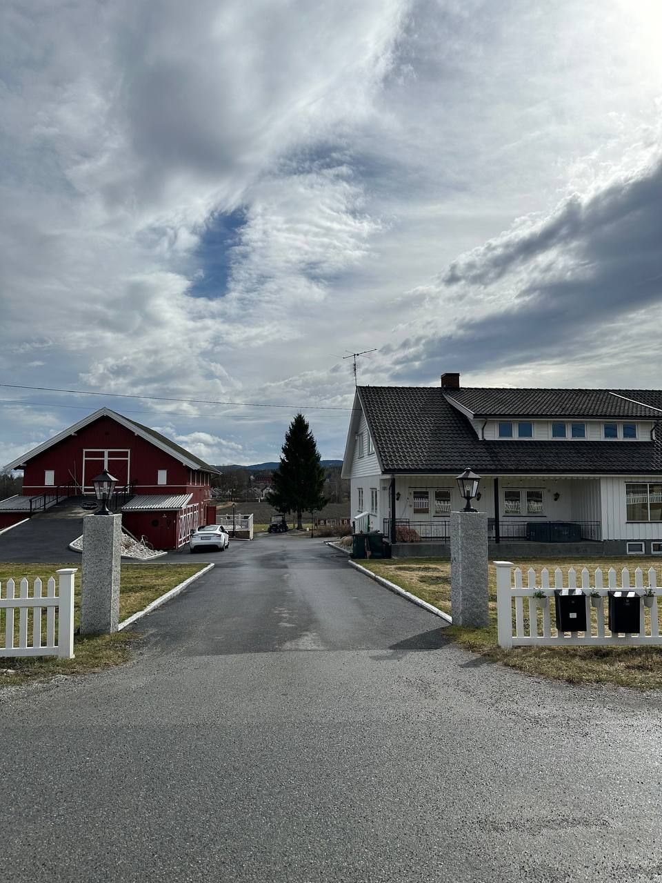 A welcoming driveway leads to two residential buildings, one painted in red and the other in a neutral tone. A white picket fence neatly outlines the property. The sky is partly cloudy, contributing to an open and spacious feel.