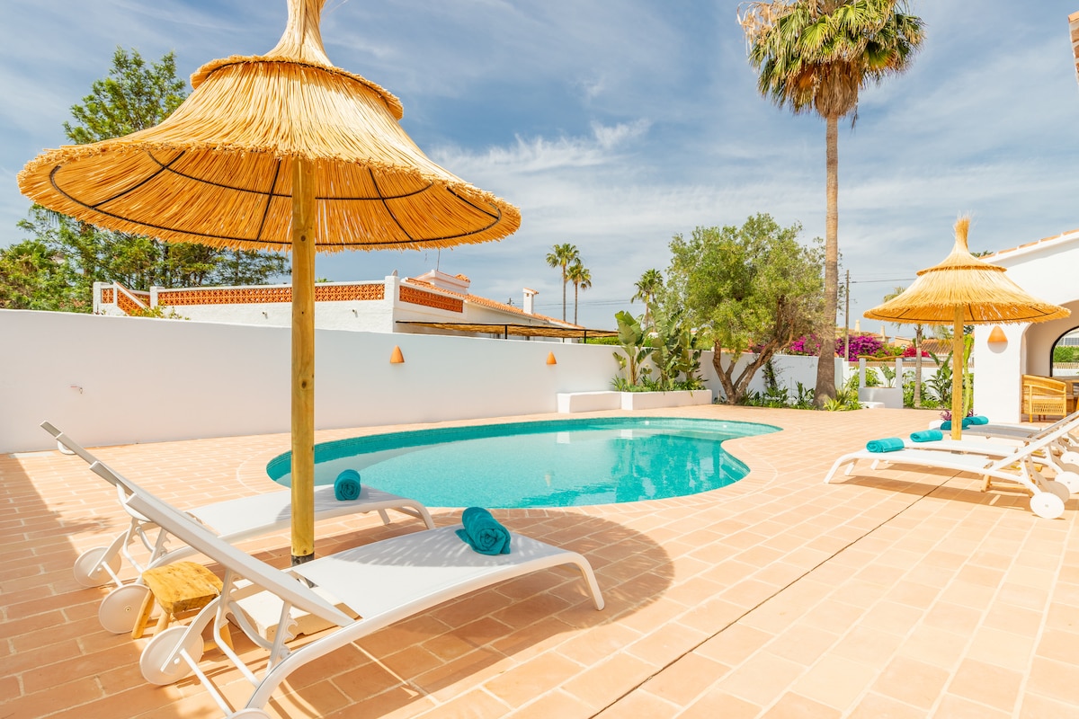 A tranquil outdoor area features a refreshing pool surrounded by a tiled patio. Two sun loungers are positioned next to the pool, and thatched umbrellas provide shade. Lush greenery and colorful flowers add vibrancy to the serene atmosphere.