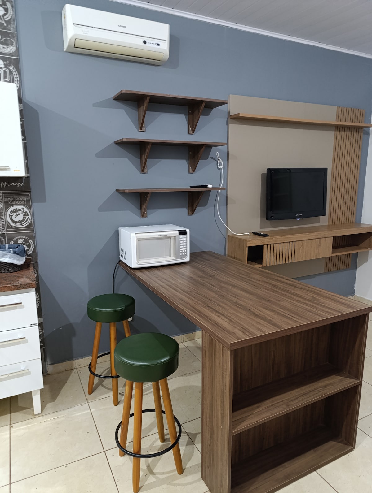 A compact kitchen area features wooden shelving above a bar-style table with two green stools. A microwave is positioned on the counter, and a wall-mounted television is visible. An air conditioning unit is installed above for climate control.