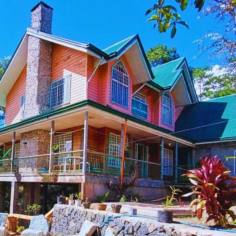 A two-story ancestral cabin features a combination of wooden and stone elements, with large windows allowing natural light to fill the interior. A green metal roof complements the pink exterior, while the surrounding greenery enhances the cabin's connection to nature.