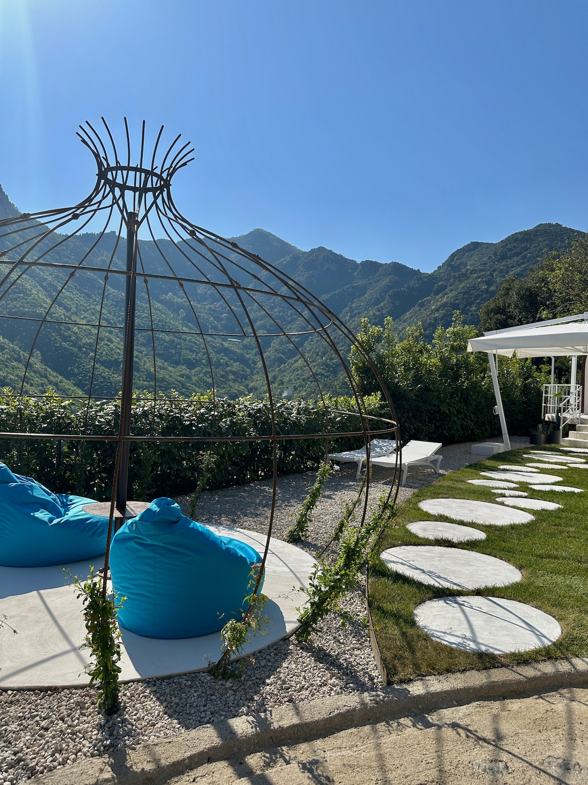 An outdoor seating area features a large, decorative structure resembling a birdcage, surrounded by vibrant green foliage. Two bright blue bean bags offer comfortable seating, while circular stone pavers lead across a grassy lawn, under a clear blue sky with mountains visible in the background.