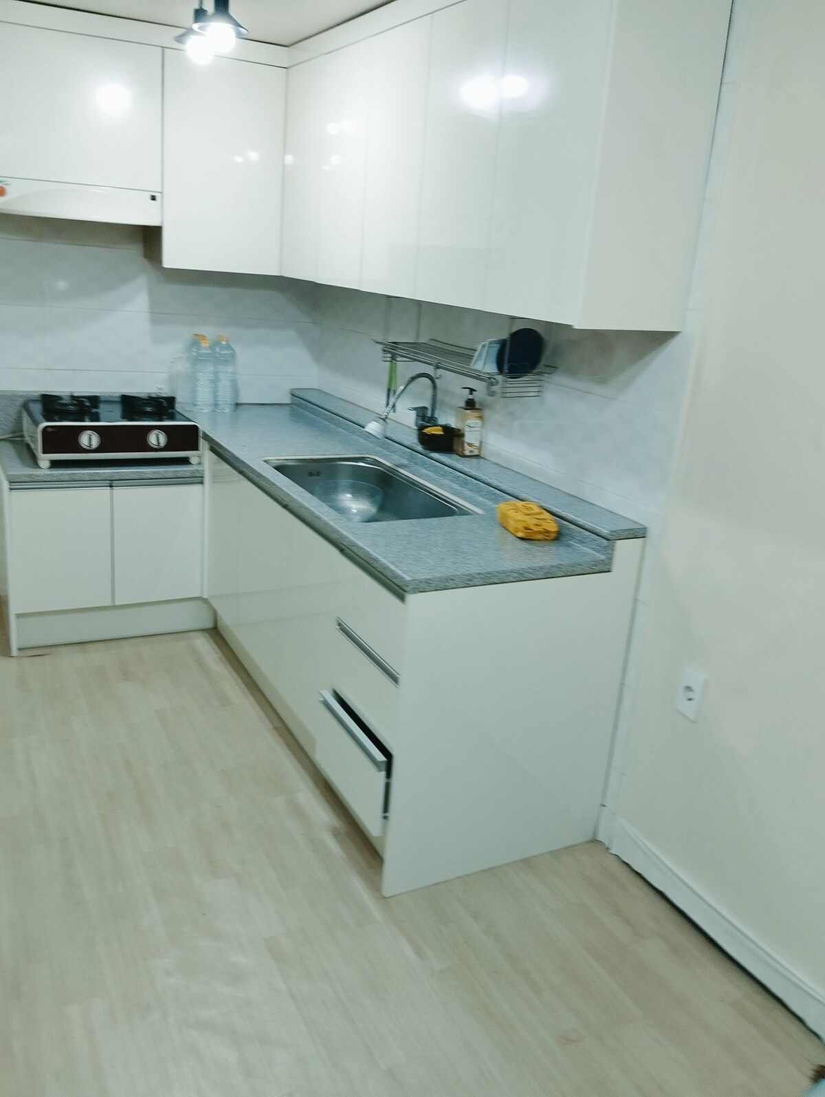 A modern kitchen is equipped with a stainless-steel sink and a sleek countertop. A gas stove is positioned beside it, with cabinets overhead providing storage. The space features a light, neutral color scheme and is illuminated by overhead lighting.