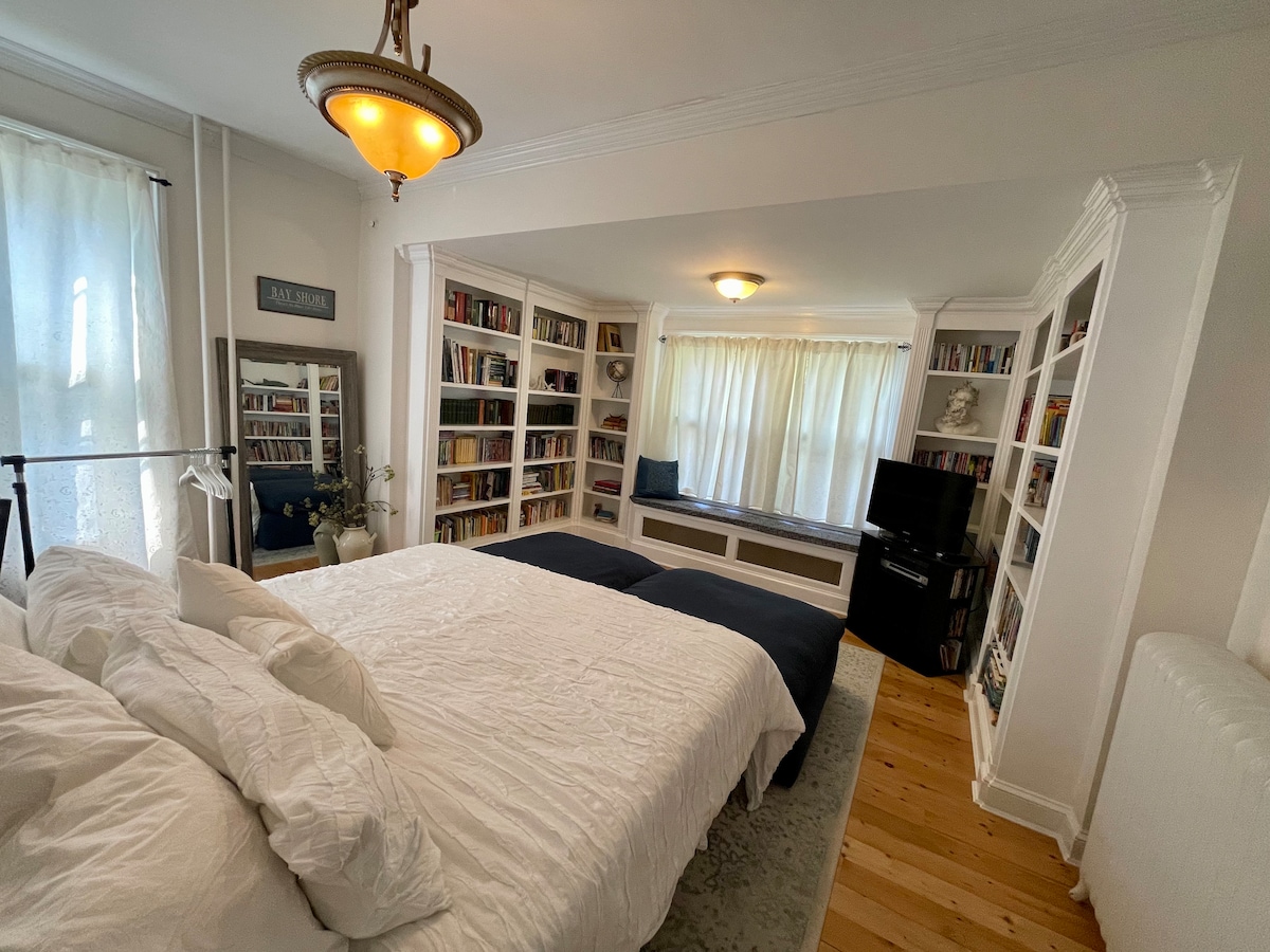 A spacious bedroom is presented with two beds side by side, surrounded by built-in bookshelves filled with books. Natural light filters through sheer curtains, illuminating the cozy nook with a window seat. A television is positioned in the corner, contributing to a welcoming atmosphere.