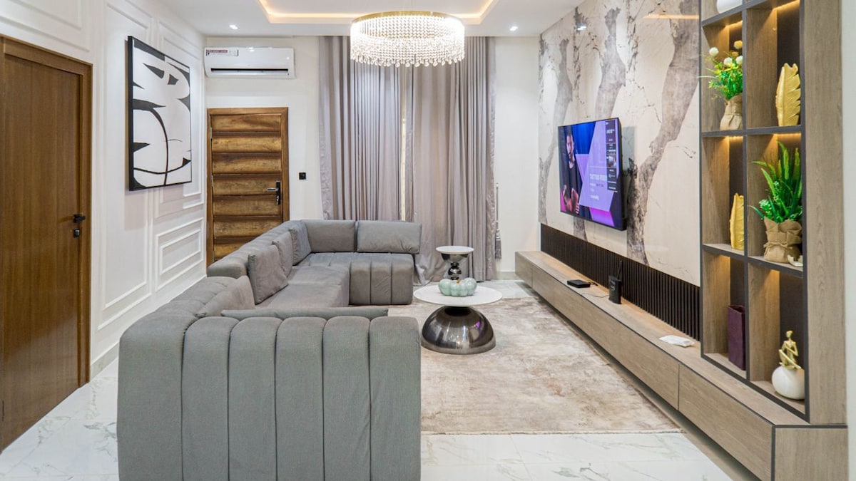 A spacious living area is furnished with a large sectional sofa and a round coffee table. A flat-screen television is mounted on a stylish media console, which features decorative plants and accents. Soft drapes cover windows that allow natural light to fill the room, complemented by modern lighting fixtures.