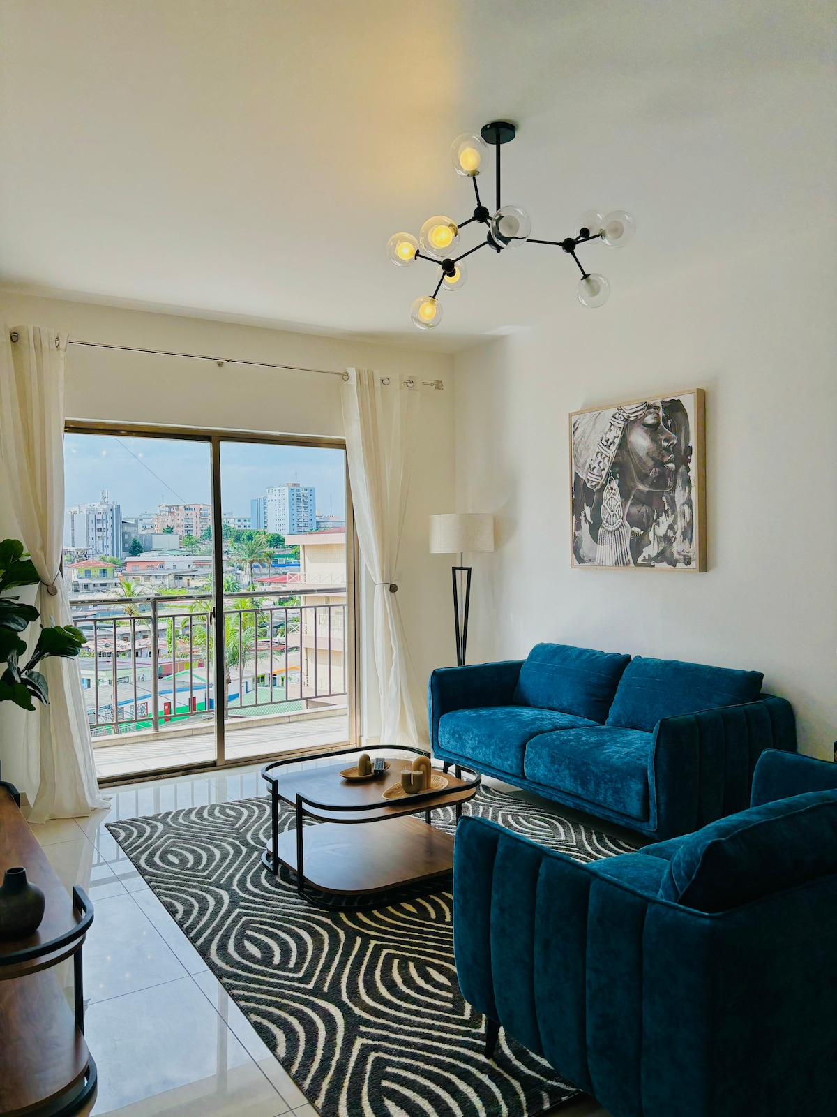 A modern living area features a plush blue sofa and a coordinating armchair, arranged around a sleek coffee table. Large glass doors allow natural light and city views, while elegant sheer curtains frame the scene. A stylish light fixture hangs above, adding a contemporary touch.