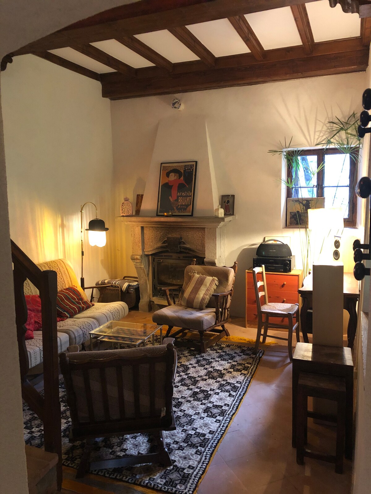 A cozy living area features warm wooden beams and a stone fireplace. A comfortable sofa is positioned with plush chairs around a patterned rug. Natural light enters through a window, highlighting a small desk and a vintage-style cabinet.