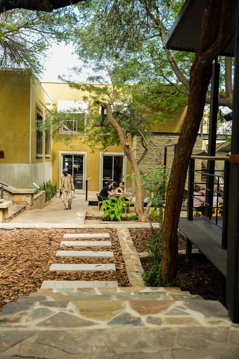 A stone pathway leads through a landscaped garden, flanked by trees and vibrant greenery. The courtyard features modern seating areas with tables, where guests can gather. The building exterior is painted in warm yellow tones, creating a welcoming atmosphere.
