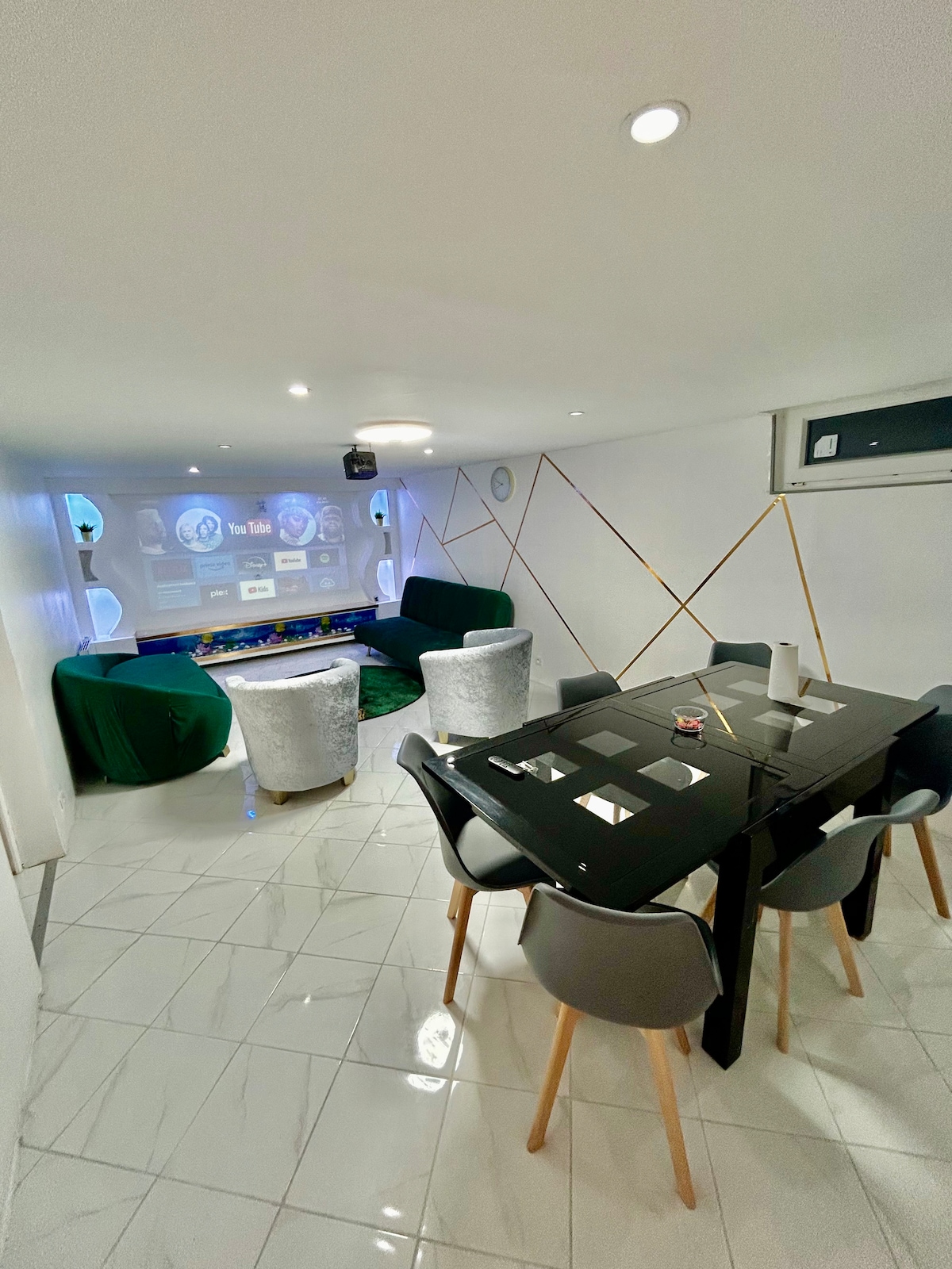 The spacious living area includes a modern dining table with six chairs and a cozy seating arrangement. A large projection screen displays multimedia content, and the room is illuminated by recessed lighting, enhancing the contemporary design. White walls complement the sleek tile floors.