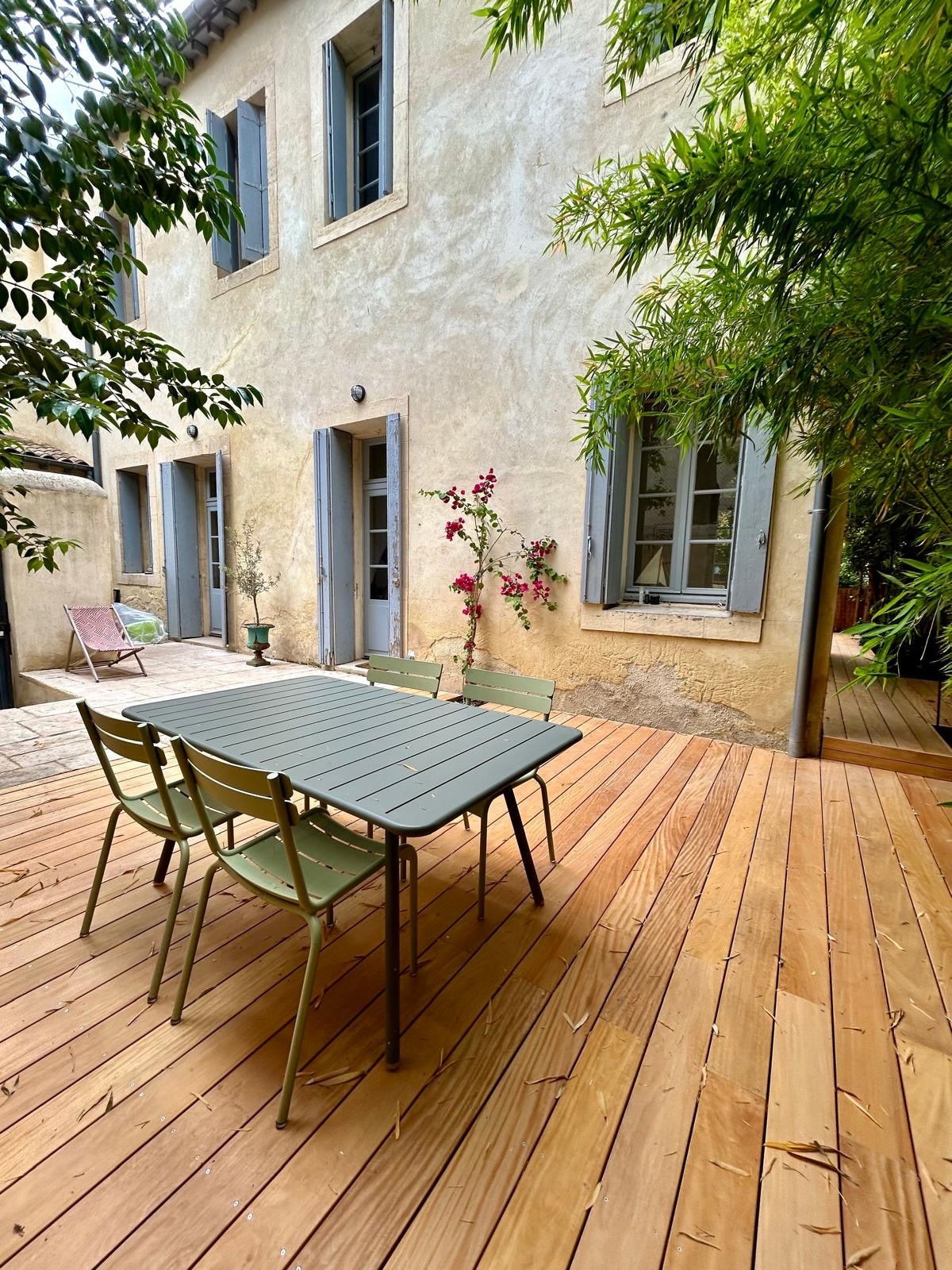 An outdoor dining area features a rectangular table surrounded by four green chairs. The warm wooden deck creates a welcoming space. Softly colored walls and greenery enhance the peaceful ambiance. Large windows provide glimpses into the interior rooms, framed by natural foliage.