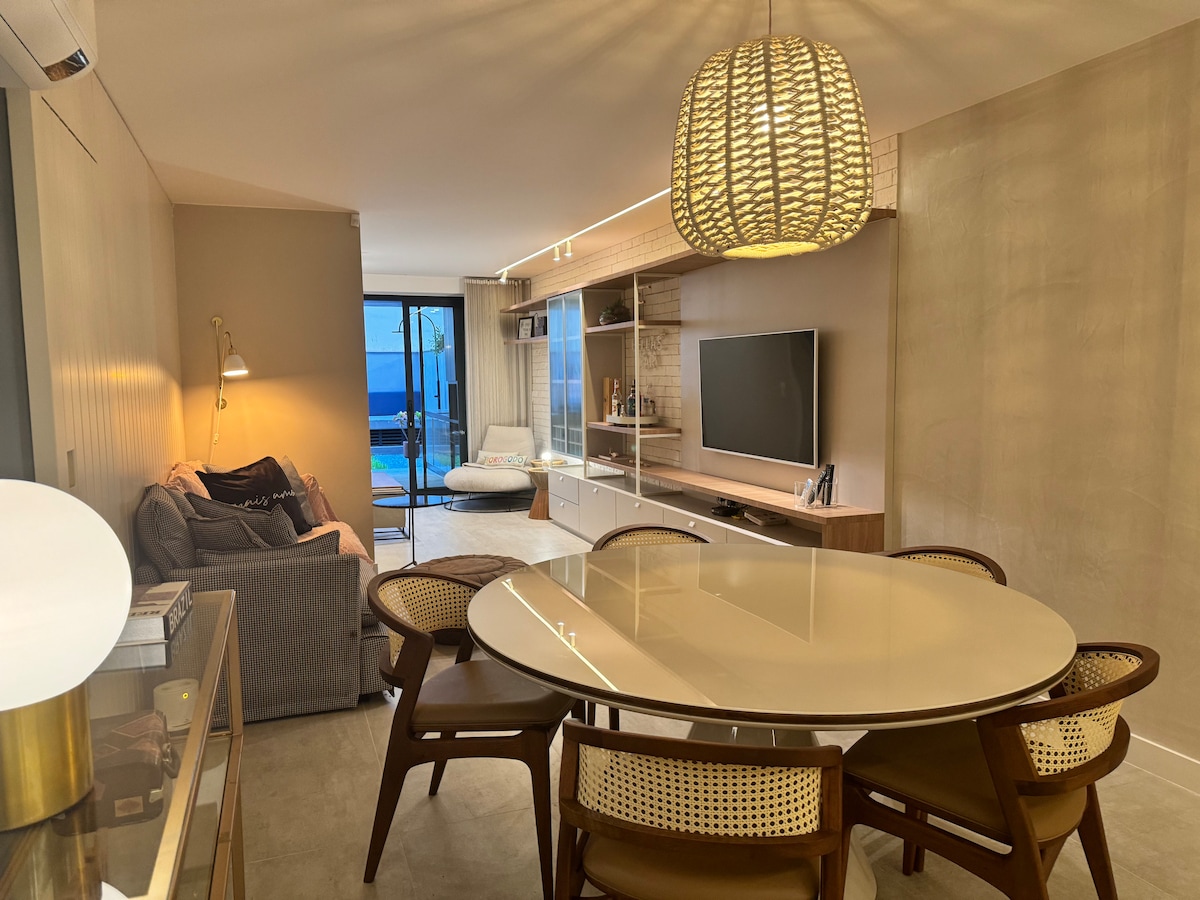 A modern living area showcases a circular dining table surrounded by elegant chairs. A comfortable sofa and a wall-mounted TV are visible, along with a stylish light fixture. Floor-to-ceiling glass doors lead to an outdoor space with scenic views.