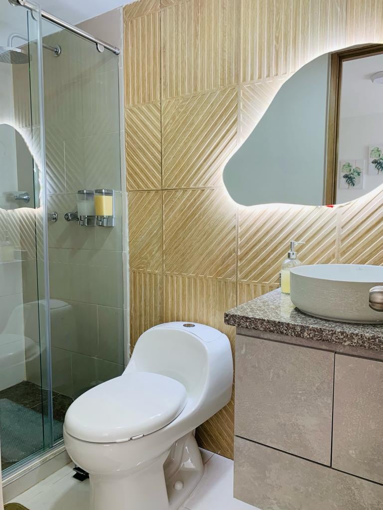 The bathroom features a modern design with textured wall tiles and a glass shower enclosure. A round sink is placed on a countertop next to a mirror that is softly illuminated. Natural light enters through a nearby window, enhancing the space's clean look.