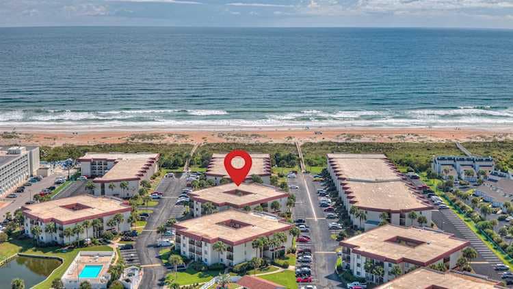 An aerial view captures the beachfront condo complex with its landscaped grounds and multiple buildings. The nearby ocean is visible, along with the sandy shoreline and gentle waves. The photo highlights the proximity of the units to the beach, emphasizing a serene coastal environment.