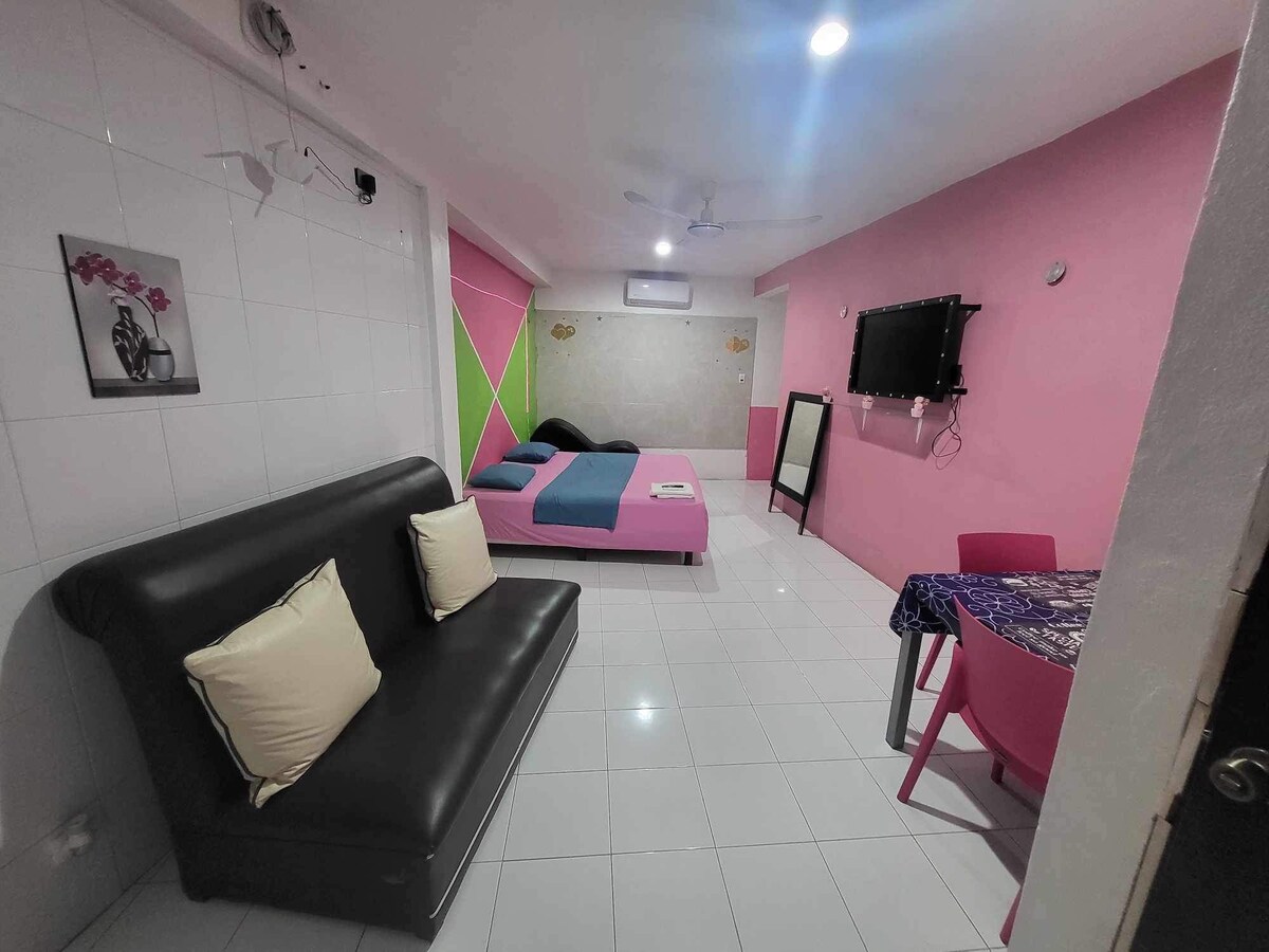 A cheerful room features a black sofa with white pillows, a spacious bed covered in blue and black linens, and a colorful accent wall with pink and green sections. A small dining area is set with a blue table and pink chairs, while a mirror and television are positioned nearby.