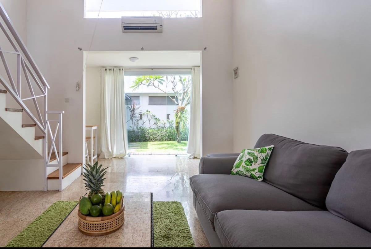 A spacious living area features a comfortable gray sofa, complemented by green decor accents. Natural light enters through large glass doors that overlook a tropical garden. A decorative basket holds fresh fruit on a low table, enhancing the inviting atmosphere.