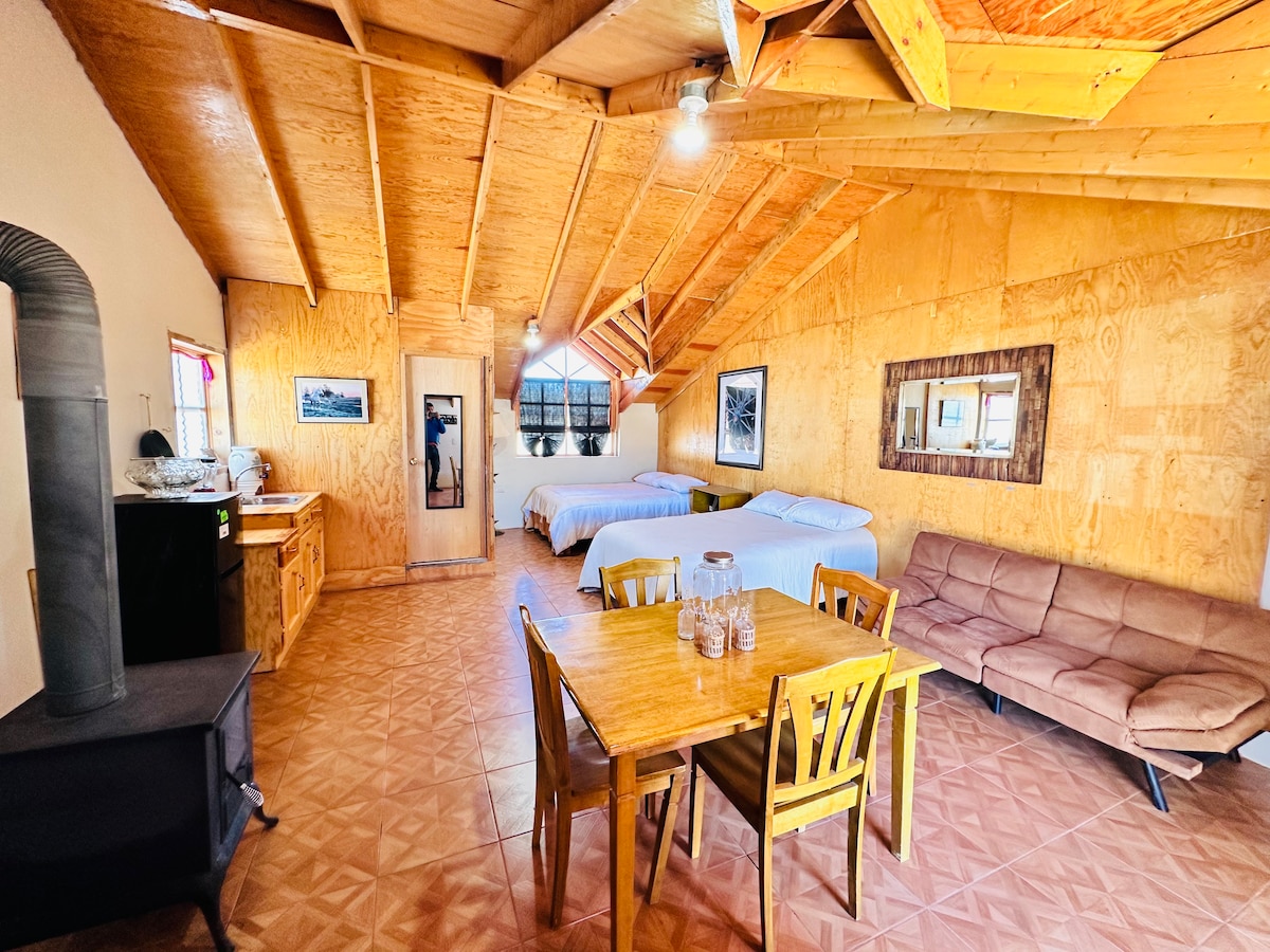 An open space features two spacious beds positioned against wooden walls, with a large window allowing natural light. A dining table with four chairs, a cozy sofa, and a stove are visible, creating a functional area enhanced by rustic wooden beams overhead.
