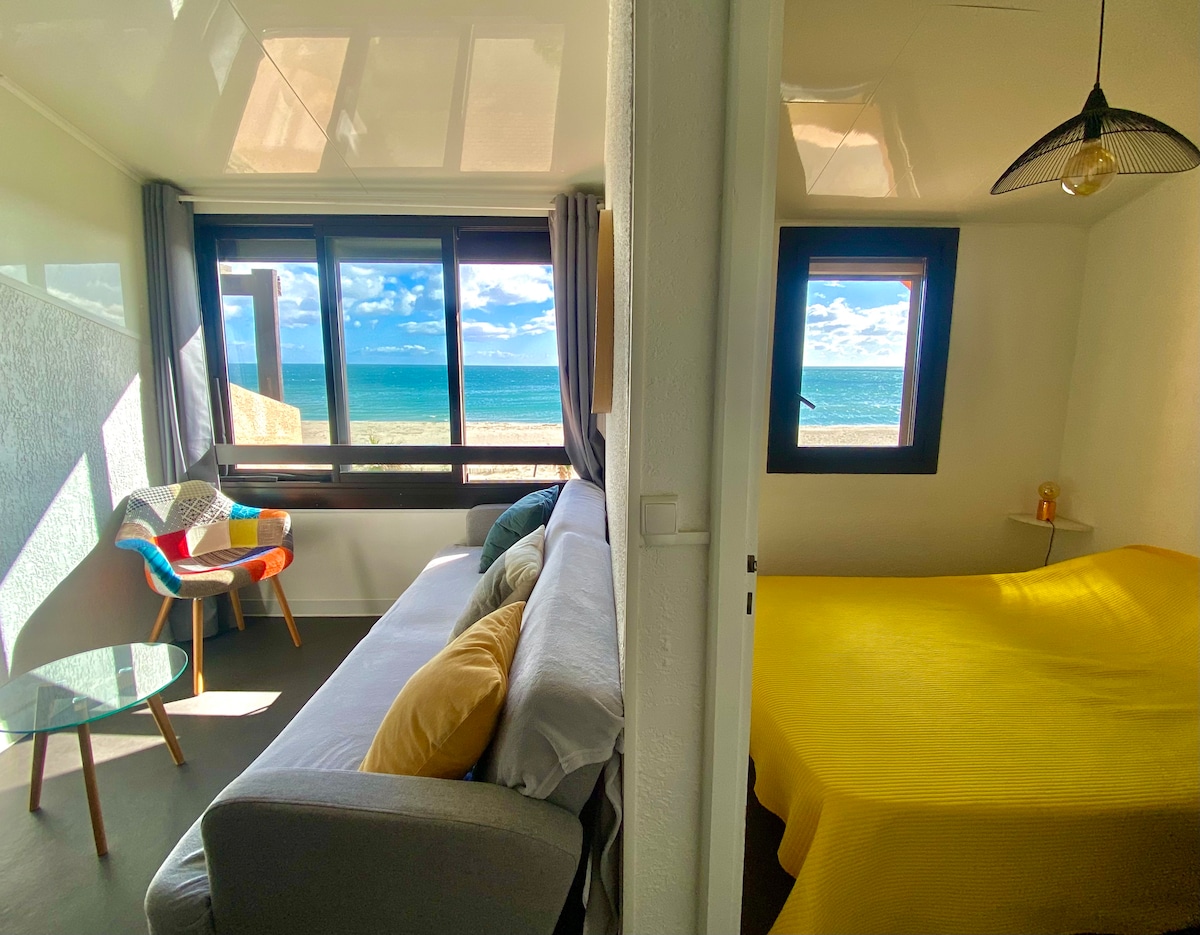 A bright and airy room features large windows with a view of the sea, providing natural light. A comfortable sofa rests beside a small glass table, while the adjoining area showcases a bed covered in a yellow blanket, creating a cozy and inviting space.