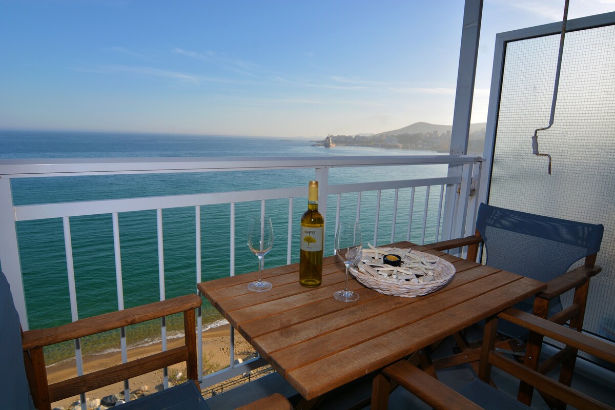 A wooden dining table is situated on the balcony, featuring a bottle of wine and two glasses. The expansive view over the sea reveals tranquil blue waters meeting a clear sky, with distant hills visible along the coastline.