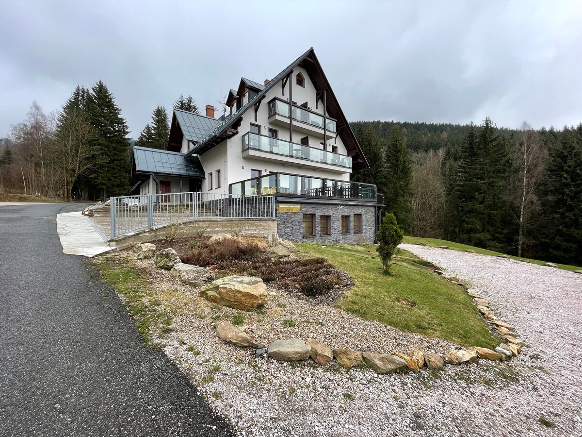 A three-story house is shown, featuring a sloped roof and a combination of stone and timber accents. Balconies extend from the upper floors, surrounded by trees. A gravel pathway leads to a landscaped area with stones and grass, enhancing the peaceful, natural setting.