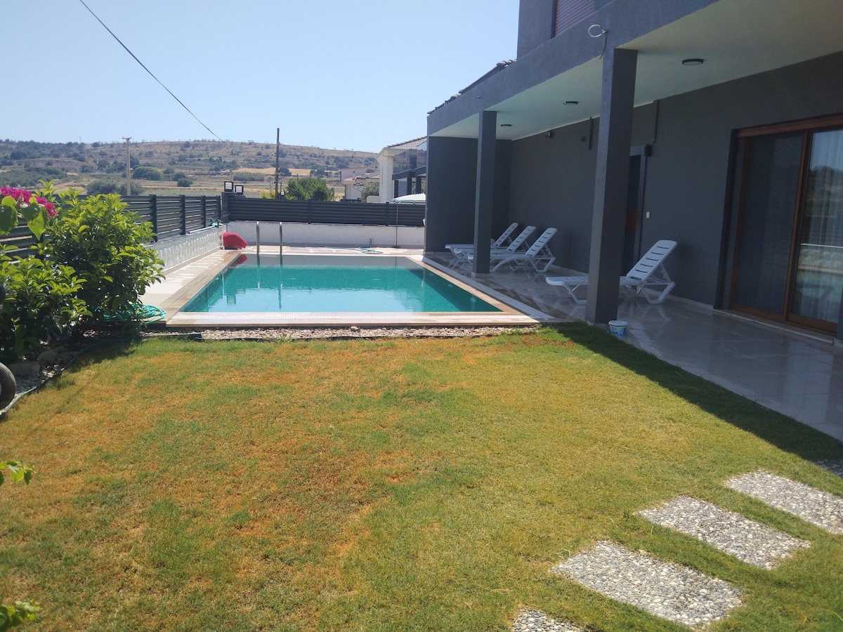 A secluded area features a private pool surrounded by well-maintained lawn. Lounge chairs are positioned by the pool, which reflects the clear blue sky. The property is framed by a modern structure and lush greenery, providing a serene outdoor setting.