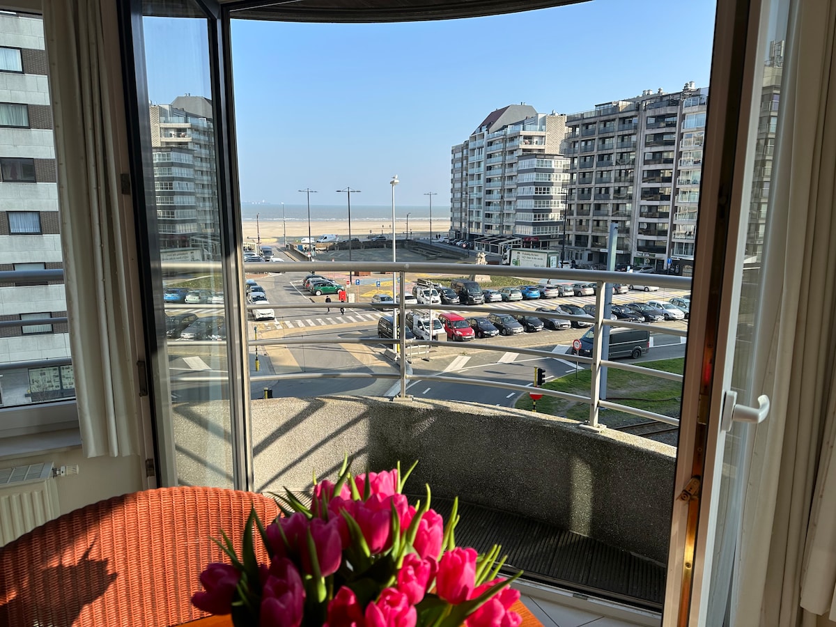A view is captured from a large window, showing a bustling street scene and nearby buildings. A flower arrangement with tulips is visible in the foreground, adding a touch of color to the scene. The coastline can be seen in the distance under a clear sky.