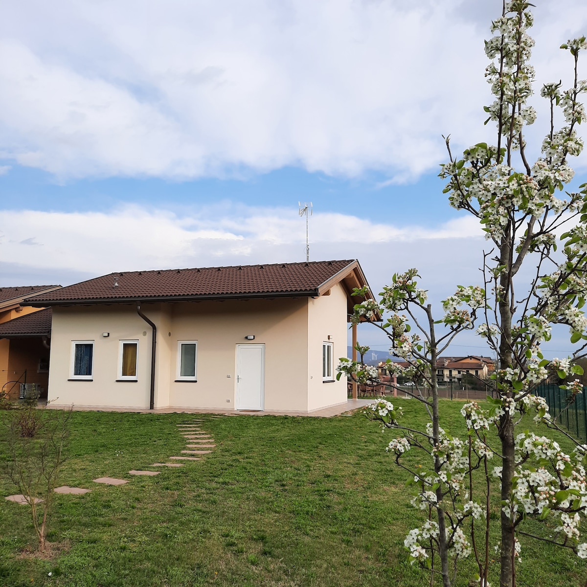 A standalone house is set within a green lawn, complemented by a flowering tree. The structure features a light-colored exterior with a sloped roof, and windows that invite natural light. A pathway of stones leads towards the front door, framed by the serene landscape.