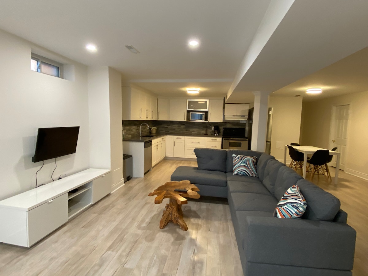 A modern living area is shown with a sectional sofa adorned with decorative pillows. A sleek television is mounted on the wall above a minimalist console. The kitchen with white cabinetry and stainless steel appliances is visible in the background, while a dining area featuring a round table is seen towards the right.