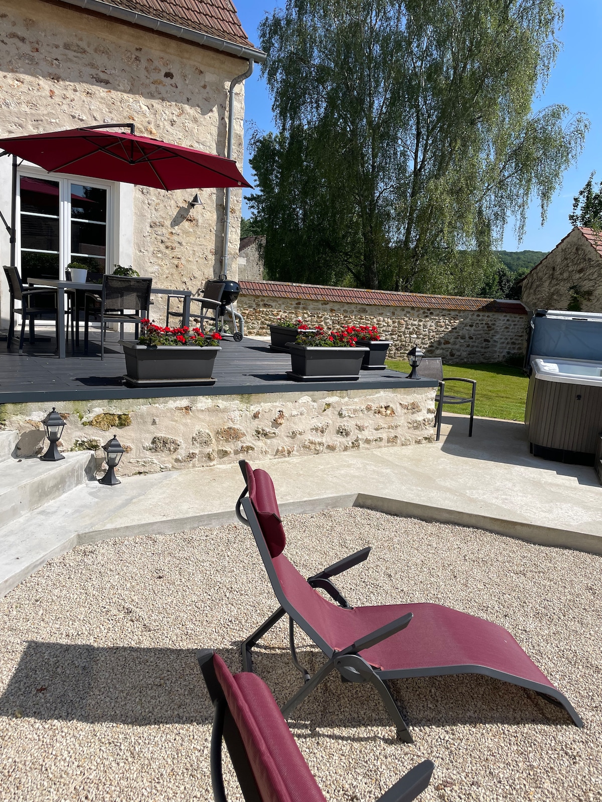 An outdoor patio area is presented, featuring comfortable lounge chairs and potted flower arrangements. A shaded dining space, marked by a large umbrella, complements the relaxing atmosphere. In the background, a hot tub is visible, surrounded by a landscaped garden.