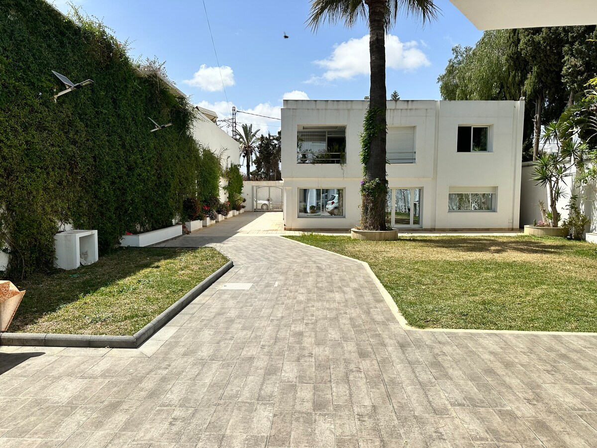 A spacious outdoor area is showcased with a neat pathway leading to a modern building. Lush greenery and palm trees frame the scene, and large windows reflect natural light. A well-maintained lawn enhances the inviting atmosphere of the property.