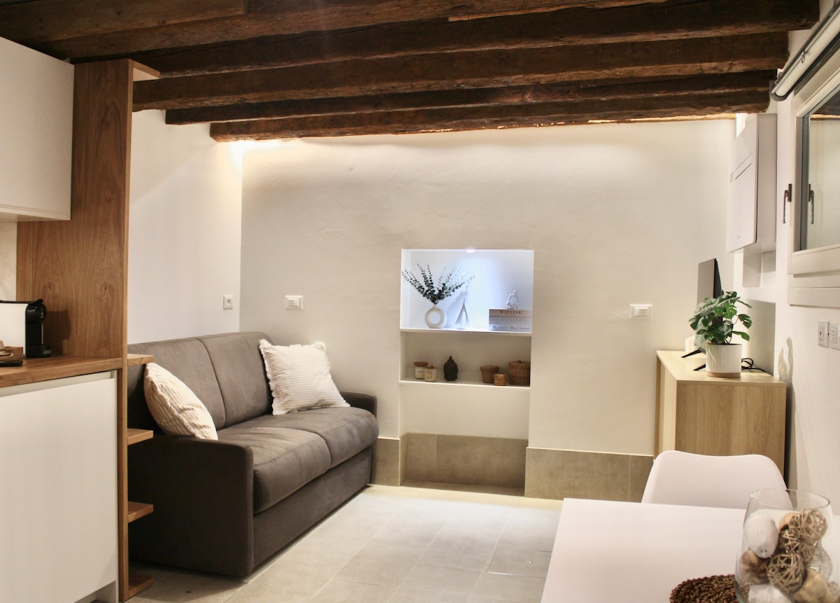 A cozy living area features a grey sofa with decorative cushions. A minimalist shelf displays plants and ceramic containers, providing a simple yet elegant touch. Soft, recessed lighting highlights the exposed wooden beams overhead, creating a warm ambiance in this inviting space.