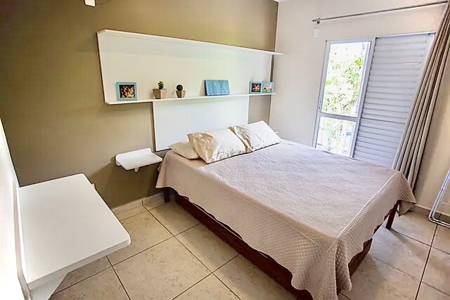 A light-filled bedroom features a spacious bed with neutral bedding, complemented by simple wall shelves displaying small decorative items. A large window provides natural light, framed by light curtains, while a desk is positioned against one wall.