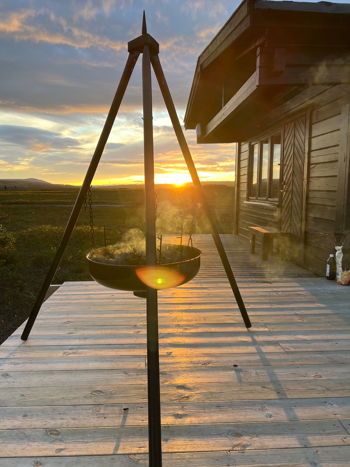 A traditional cooking tripod stands on a wooden deck, with a pot hanging above a gentle flame. The sunset creates a warm glow in the background, enhancing the serene landscape of mountains and open fields.