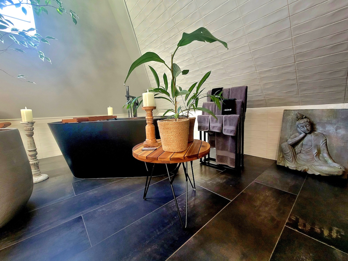 A serene spa-inspired bathroom is showcased, featuring a modern soaking tub and a minimalist design. A potted plant sits on a round wooden table, complemented by elegant candles and soft towels arranged nearby. A Buddha statue provides a calming focal point, enhancing the tranquil ambiance.