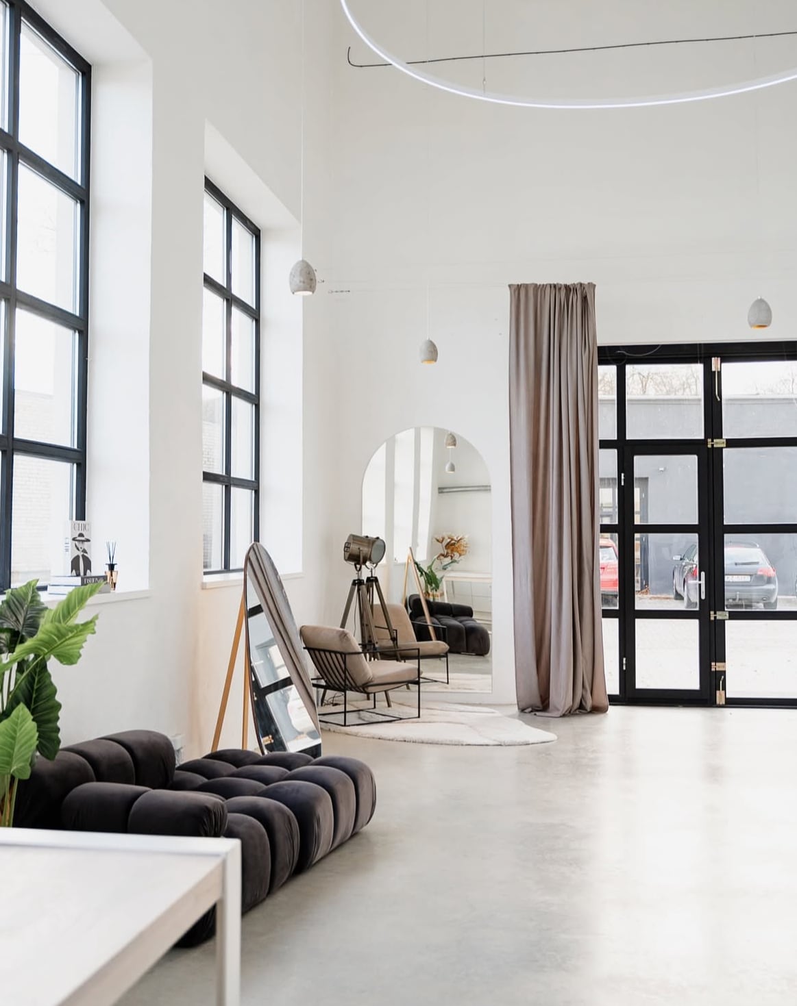 A spacious room features high ceilings and large windows allowing natural light to flood in. A dark sofa sits opposite two armchairs, with a mirror leaning against the wall. Elegant curtains frame a doorway, while decorative plants add a touch of greenery.