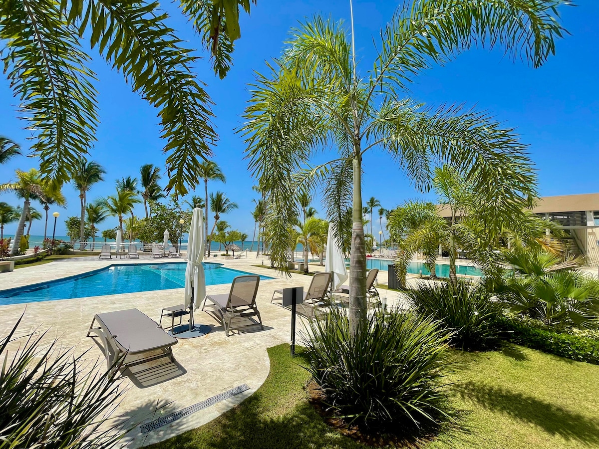 A serene pool area is surrounded by lush palm trees and neatly trimmed shrubs. Lounge chairs are positioned near the water's edge, inviting relaxation. The clear blue sky enhances the tranquil atmosphere, while the ocean is visible in the background.