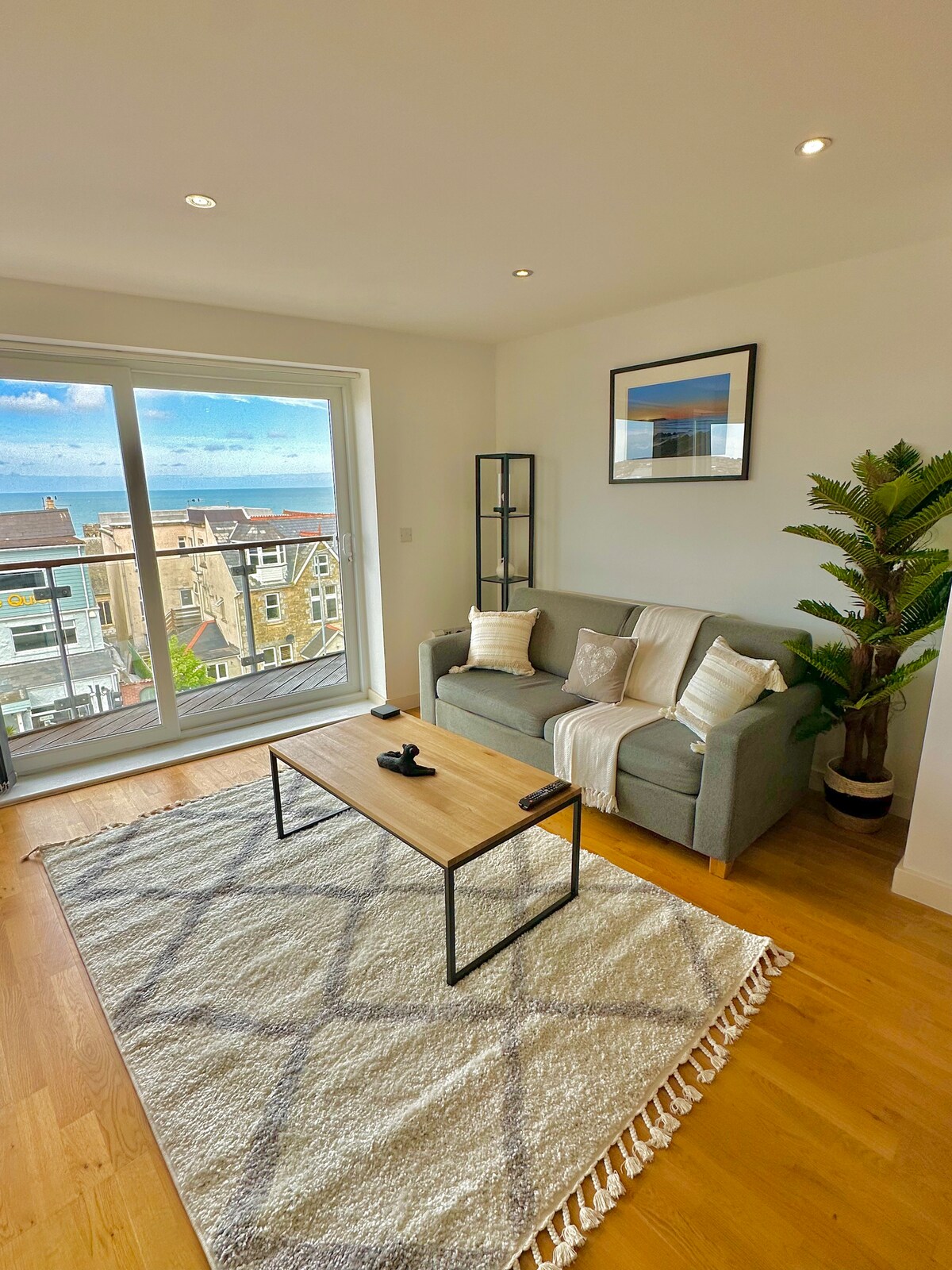 A modern living area features a comfortable grey sofa adorned with light cushions. A simple wooden coffee table sits on a textured area rug, while a tall plant adds greenery to the space. Large glass doors provide a view of the ocean and town.