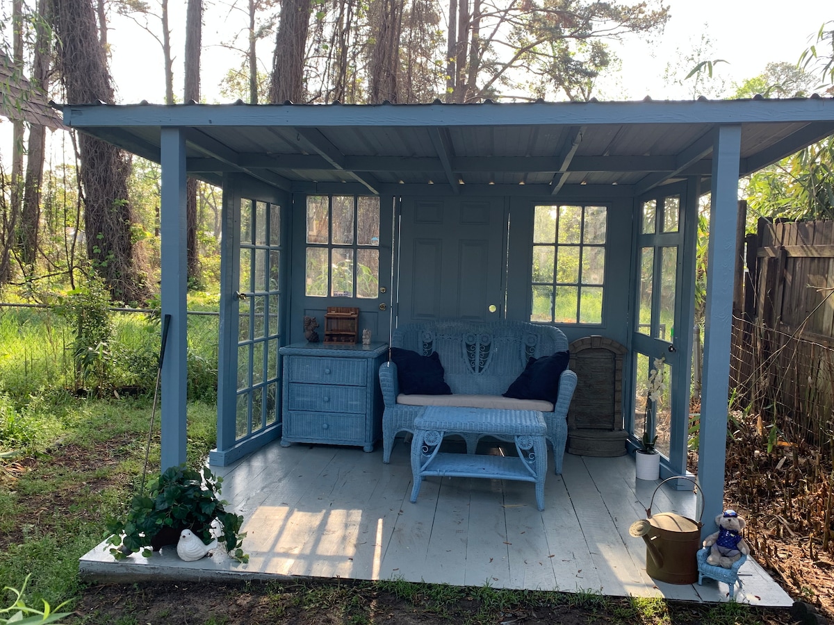 A charming outdoor seating area is showcased under a shaded structure with a light blue color. It features a wicker-style bench and a small table, surrounded by large windows that provide views of the lush greenery beyond. Decorative elements are placed throughout the space.