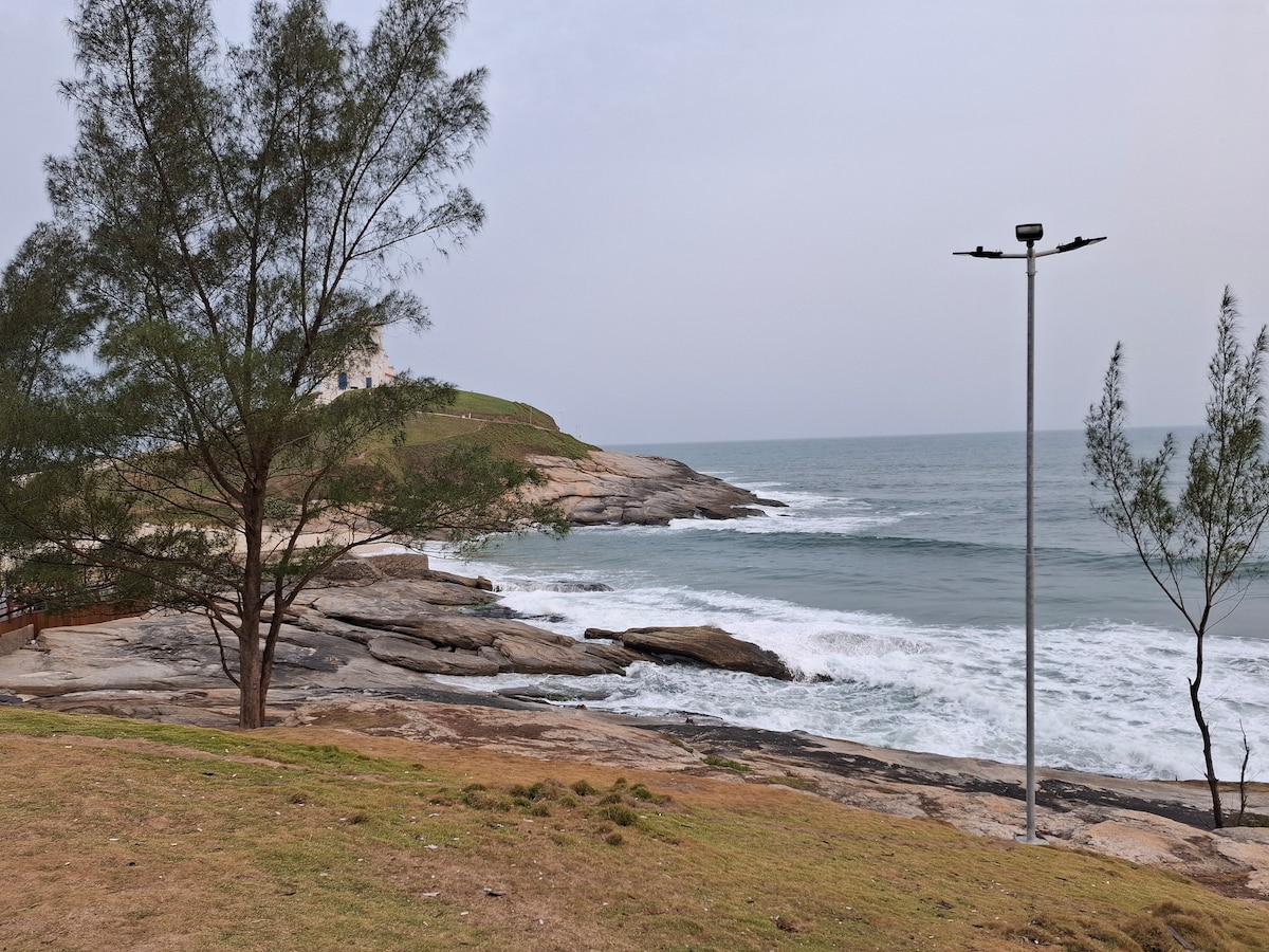 A serene coastal view is captured, showcasing gentle waves lapping against rocky shores. Tall trees frame the scene, while a lamppost stands along the path. The soft contours of the landscape lead up to lush grassy areas, with hints of distant land visible across the water.