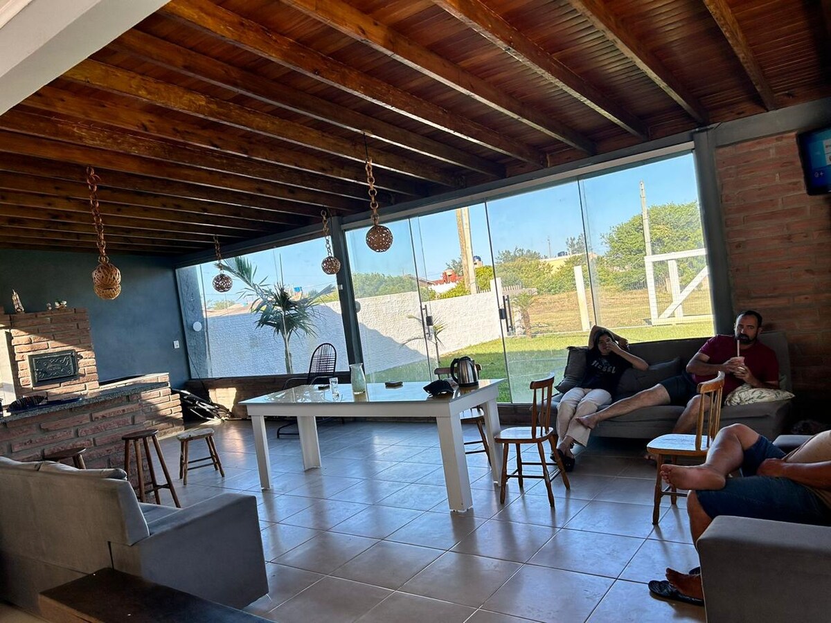 An open and airy living area features wooden beams and large windows, allowing ample natural light. A sleek white table sits at the center, surrounded by wooden chairs. A stone fireplace and cozy seating options enhance the space, with outdoor views visible through the glass.
